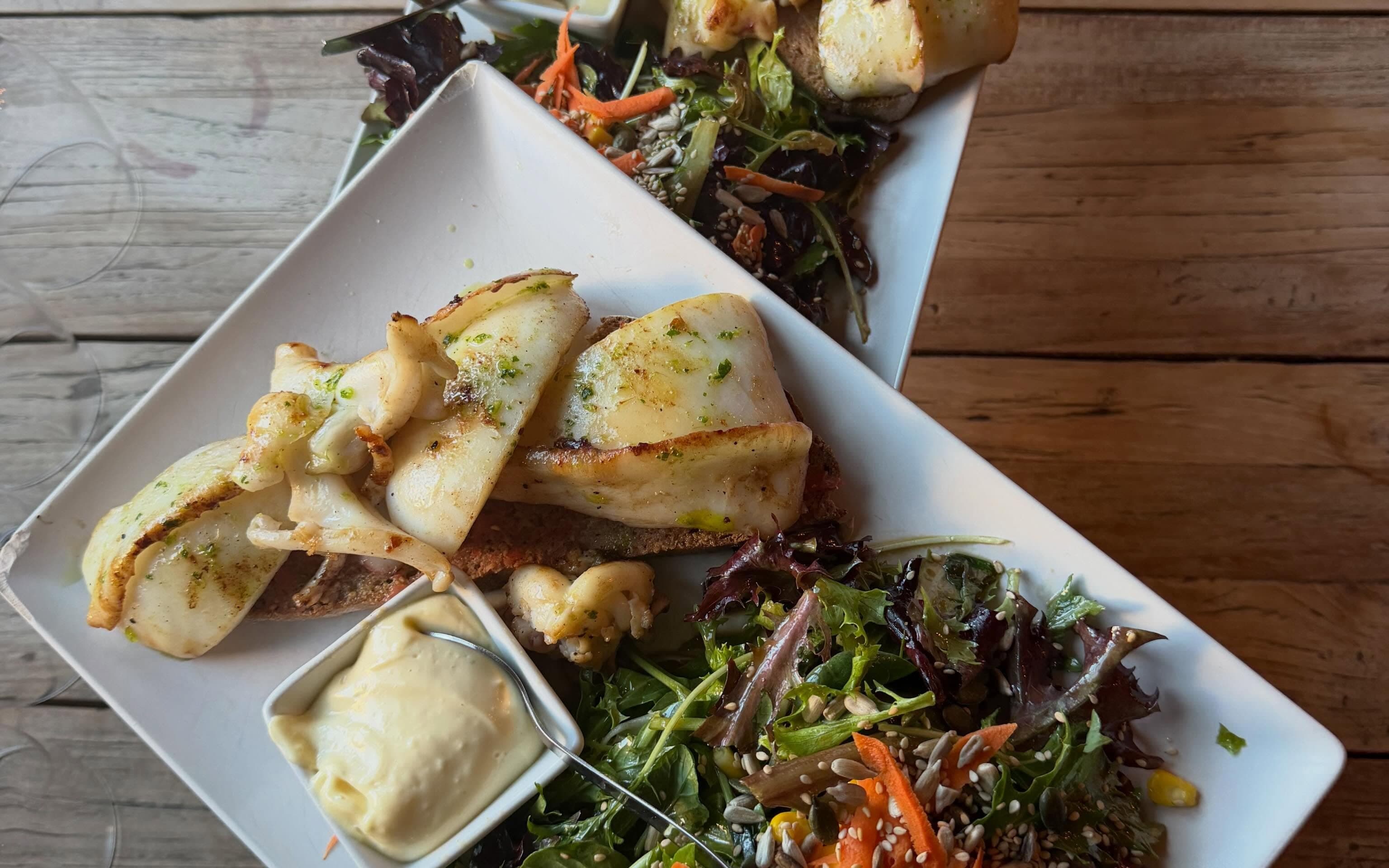 Dos platos de sepia a la plancha con pa amb oli, alioli y ensalada, sobre una mesa de madera