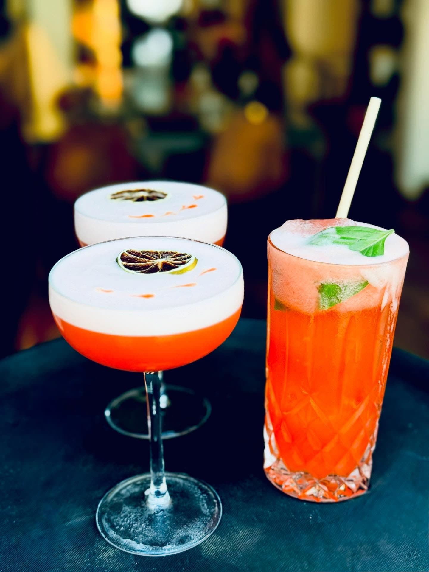 Dos cócteles con espuma y un vaso alto, con luces cálidas detrás